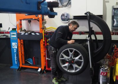 In der Blochberger Werkstatt in Prutz prüft ein Mechaniker ein Autorad auf einer Auswuchtmaschine. Der saubere Werkstattboden und die ordentlich angeordneten Werkzeuge werden durch große Fenster im Hintergrund beleuchtet.