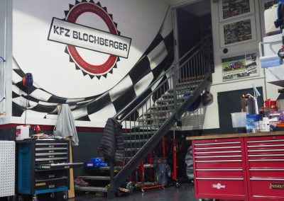 Ein Werkstatt-Interieur in Prutz mit Werkzeugschränken, hängenden Mänteln, einer Treppe und einem Wandbild mit der Aufschrift „KFZ Blochberger“ über einer schwarz-weiß karierten Flagge. Verschiedene Werkzeuge und Geräte sind zu sehen.