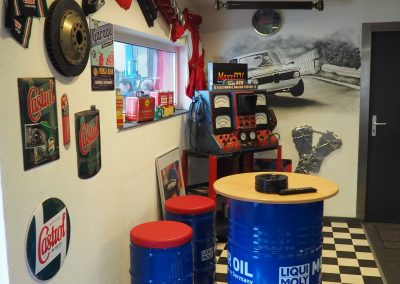 Ein Werkstattraum im Garagen-Stil mit kariertem Boden, Wanddeko mit Automotiv und Vintage-Schildern. Blaue Ölfässer dienen als Hocker und Tisch. Es gibt ein Fenster, verschiedene Auto-Erinnerungsstücke und ein Arcade-Spiel mit Rennsport-Thema.